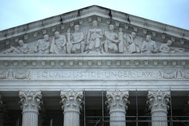 The roofline with carvings on it of a large, white, pillared building.