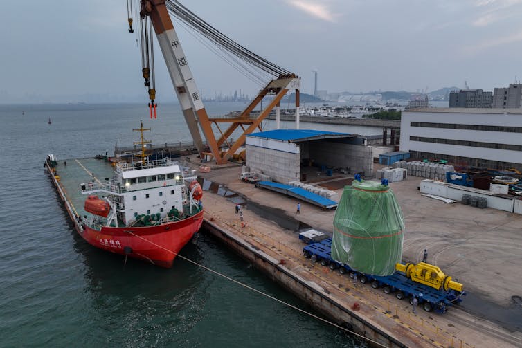 A large green item is on a pier next to a ship and a crane.